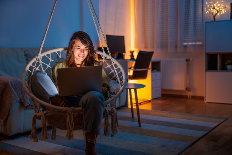 Smiling woman working on a laptop in a cozy, dimly lit room at night.